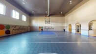 The Timberline Place indoor sports facility is a generously sized, single-level court designed to accommodate a variety of athletic activities. The court surface features a durable interlocking tile floor in a two-tone grey and blue color scheme, with clearly marked regulation lines for basketball and additional multi-sport configurations. Two basketball hoops with glass backboards are mounted at either end of the court, and a portable net post stands ready for volleyball or similar net sports.

The soaring ceiling height allows ample vertical clearance for gameplay, and the space is illuminated by a combination of overhead track lighting and natural light streaming through windows positioned high along the upper walls. A wall-mounted electronic scoreboard displaying home and visitor scores adds a competitive, professional atmosphere to the space.

Padded wall panels line the lower portion of the perimeter walls, providing both safety and acoustic management. The Timberline Place branding is prominently displayed across the main wall in a bold, stylized graphic featuring the community's colorful logo. An arched architectural detail near one end wall adds a subtle decorative element to the otherwise athletic-focused interior. The overall ambiance is clean, functional, and well-equipped for both organized competition and recreational play.