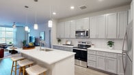 This single-floor, 2-bedroom, 2-bathroom residence offers a clean, modern aesthetic throughout. The open-concept kitchen and living area serves as the heart of the home, featuring a large center island with a built-in sink, white quartz countertops, gray shaker-style cabinetry, a white subway tile backsplash, and stainless steel appliances including a range, microwave, and refrigerator. Pendant lighting above the island adds a stylish touch, while light wood-look flooring flows seamlessly throughout the main living spaces. The living room benefits from natural light through a full-length door and transom window, lending an airy, open feel to the space. The generously sized primary bedroom includes a ceiling fan, dedicated workspace area, and direct access to a walk-in closet equipped with extensive wire shelving systems. The second bedroom is bright and welcoming, with a large window dressed in sheer curtains. Both bathrooms are finished with clean, contemporary tilework; one features a full soaking tub and tiled surround. A dedicated laundry room houses a full-sized top-load washer and dryer with overhead wire shelving for added storage. Two closets throughout the residence provide ample organizational space.