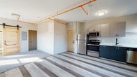 This single-floor unit delivers a compelling industrial-modern aesthetic, where raw architectural elements meet contemporary finishes. The open living area is defined by striking exposed brick walls that add warmth and texture, while bold orange overhead piping runs across the white ceiling, reinforcing the loft-inspired character. Multi-toned luxury vinyl plank flooring in varying shades of grey flows throughout the space, creating a dynamic visual contrast underfoot. Natural light filters in through white-framed windows, brightening the interior and highlighting a rustic barn-style sliding door that connects living spaces with a charming, functional touch. The kitchen is well-appointed with two-toned cabinetry — dark navy lower cabinets paired with light grey uppers — complemented by stainless steel appliances including a range, microwave, refrigerator, and dishwasher. A matte black faucet over a single-basin sink adds a refined modern detail. A mini-split HVAC unit provides individual climate control. The bathroom features a glass-enclosed walk-in shower with large-format tile, a white vanity, and a dark-framed mirror, completing the space with a clean, polished finish.