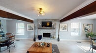 This single-floor space offers an open, flowing layout that seamlessly connects the living area, dining space, and bar area. The living room serves as the focal point, anchored by a floor-to-ceiling stacked-stone fireplace with a rustic wood mantel and wall-mounted television flanked by candle sconces. Bold, dark-stained wood beams span the ceiling overhead, contrasting beautifully against the textured white ceiling and soft blue-gray walls. Light wood-style flooring runs throughout, lending a clean, cohesive look to the entire space. A semi-flush brass and glass light fixture adds a warm, stylish glow. Multiple windows along the perimeter allow generous natural light to filter in, creating a bright and welcoming atmosphere. The dining area accommodates a round table and chairs beneath a matching brass fixture, with large windows offering views of the surrounding greenery. A cozy secondary dining nook sits adjacent to a stacked-stone bar counter with pendant lighting, adding a casual entertainment element. Crown molding and consistent finishes throughout the space tie each zone together, creating a polished yet relaxed ambiance ideal for everyday living and entertaining alike.