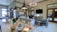 This single-floor communal lounge is thoughtfully designed to offer a comfortable and inviting atmosphere for gathering and leisure. The space features notably tall ceilings that enhance the sense of openness, complemented by a sophisticated mix of lighting fixtures, including an industrial-style pendant lamp, a classic chandelier, and a contemporary floor lamp, each contributing to a warm and layered ambiance.

A large granite-topped counter anchors the foreground, serving as a casual refreshment and social hub, currently stocked with snacks and beverages. The seating area is furnished with neutral-toned sofas and armchairs arranged around a central rug, creating an intimate yet spacious lounge setting. A wall-mounted flat-screen television adds an entertainment focal point to the space.

Decorative orange-accented wall art adds a pop of color against the gray and beige walls, while a rustic wooden console and textured décor elements lend a transitional aesthetic. A glass-paneled door at the rear draws in natural light and hints at an adjacent room or outdoor area beyond. Workstation setups are visible along one wall, adding a functional, multi-use dimension to the lounge.