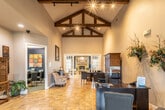 This single-floor community space combines rustic architectural charm with functional, well-appointed areas designed to welcome and serve visitors. The main reception area immediately impresses with its soaring vaulted ceiling accented by rich, dark-stained exposed wood beams and track lighting that illuminates the warm terracotta tile flooring below. Dark-finished workstations provide a professional setting for staff, while a large wooden armoire adds a classic, homey touch to the space.

Beyond the reception area, French doors open into a spacious lounge that serves as the heart of the facility. A dramatic floor-to-ceiling stone fireplace anchors the room, flanked by pairs of French doors that flood the interior with natural light and offer views to the outdoors. Deep navy accent walls, comfortable seating arrangements, and a rustic wooden coffee table create an inviting and relaxed atmosphere.

A separate office or meeting room off the main hall provides a quieter workspace, while an adjacent kitchenette area features white cabinetry, a dark countertop breakfast bar with wrought-iron barstools, and decorative wall accents. A media area with a wall-mounted television and dining table rounds out the space, making it well-suited for both social gatherings and day-to-day operations.