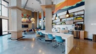 Located at 2125 E Apache Blvd in Tempe, Arizona, this dynamic single-floor community amenity space offers a generously sized, open-concept layout designed for social gathering and relaxation. The space is defined by soaring double-height ceilings in the main lounge area, allowing abundant natural light to pour in through expansive floor-to-ceiling windows and glass doors that open directly to an outdoor patio. Polished concrete floors reflect light throughout, reinforcing the space's airy, contemporary feel.

Bold, colorful murals — including a sweeping desert-inspired landscape above the kitchenette and a vibrant botanical artwork in the lounge — serve as striking focal points. The kitchenette features open wood shelving, white tile backsplash, and a long marble-topped island surrounded by teal seating, ideal for casual dining or socializing. Pendant lighting in varied styles adds warmth and visual interest throughout.

The lounge area offers multiple seating arrangements, including a large cognac leather sectional positioned beside a graphite accent wall with an uplifting typographic mural. Mid-century modern accent chairs, patterned area rugs, and wood-paneled walls with decorative lattice screens contribute to the space's layered, design-forward character. A pool table anchors the recreation zone, while a wall-mounted TV provides additional entertainment. Concrete structural columns and exposed ductwork add an industrial edge that balances the space's warm, eclectic aesthetic.