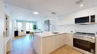 This contemporary 1-bedroom, 2-bathroom residence at 95 Anderson Street in Hackensack, NJ offers a well-designed, modern living experience. The open-concept kitchen and living area serves as the heart of the home, featuring sleek white flat-front cabinetry, a quartz waterfall peninsula with bar seating, stainless steel appliances including a gas range and French door refrigerator, and a textured tile backsplash. Warm hardwood flooring flows throughout the main living areas, complementing the bright, white palette. The living room is bathed in natural light through large floor-to-ceiling windows that frame neighborhood views, creating an airy and inviting atmosphere. A floating media console and generous wall space add to the clean, minimalist aesthetic. The primary bedroom is a comfortable retreat with a sizable window offering outdoor views, hardwood flooring, and direct access to a well-appointed en-suite bathroom. The bathrooms are finished with large-format white tile, warm wood vanities, and matte black fixtures, striking a balance between modern and organic design. Throughout the residence, crisp white walls, recessed and pendant lighting, and thoughtful layout choices combine to create a cohesive, contemporary living space.