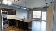 This single-floor space showcases a thoughtfully designed open-concept layout that seamlessly blends modern and industrial aesthetics. The kitchen serves as a central feature, boasting a large granite-topped island with an undermount sink, dark espresso cabinetry, and stainless steel appliances including a dishwasher. Stylish pendant lights hang above the island, complementing the exposed ductwork that runs along the ceiling — a nod to the space's industrial character. Rich, dark hardwood flooring extends throughout, adding warmth and contrast to the cool gray walls. The kitchen flows naturally into the living area, creating a spacious, connected environment ideal for both everyday living and entertaining. Large windows near the living area allow abundant natural light to fill the space, enhancing its open and airy feel. A door adjacent to the windows provides additional access and further brightens the interior. The combination of high ceilings with visible mechanical elements, contemporary finishes, and an open floor plan gives this space a distinctive urban loft character that is both functional and visually striking.