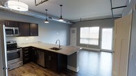 This single-floor space showcases a thoughtfully designed open-concept layout that seamlessly blends modern and industrial aesthetics. The kitchen serves as a central feature, boasting a large granite-topped island with an undermount sink, dark espresso cabinetry, and stainless steel appliances including a dishwasher. Stylish pendant lights hang above the island, complementing the exposed ductwork that runs along the ceiling — a nod to the space's industrial character. Rich, dark hardwood flooring extends throughout, adding warmth and contrast to the cool gray walls. The kitchen flows naturally into the living area, creating a spacious, connected environment ideal for both everyday living and entertaining. Large windows near the living area allow abundant natural light to fill the space, enhancing its open and airy feel. A door adjacent to the windows provides additional access and further brightens the interior. The combination of high ceilings with visible mechanical elements, contemporary finishes, and an open floor plan gives this space a distinctive urban loft character that is both functional and visually striking.