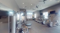 This single-floor fitness center is designed to offer a comprehensive workout experience in a clean, well-organized environment. The space features a neutral color palette of soft grays and warm taupes, contributing to a calm yet motivating atmosphere. Recessed lighting is installed throughout the ceiling, complemented by multiple ceiling fans that promote air circulation during workouts. Large windows along the perimeter allow generous amounts of natural light to flood the interior, creating an open and energizing ambiance.

The strength training area is outfitted with a multi-functional cable machine and a dedicated free weights section, including adjustable benches and a variety of dumbbells suited for diverse training regimens. On the opposite side of the space, a row of treadmills is neatly arranged along the wall, each positioned near windows to allow users to enjoy natural light during cardio sessions. A wall-mounted flat-screen television is also present, providing entertainment for those using the cardio equipment.

Additional fitness accessories, including stability balls and other training aids, are visible near the windows, rounding out the space's versatility. The flooring is finished with durable, low-pile carpet suited for a fitness environment. Overall, this fitness center delivers a well-rounded facility with thoughtful design and a broad selection of equipment.