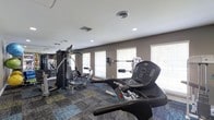 The fitness center is a thoughtfully designed single-level space built to support a well-rounded workout routine. The room is lined with windows fitted with horizontal blinds, allowing natural light to fill the space while maintaining privacy. Recessed lighting in the white ceiling supplements the natural light, ensuring the area remains bright and functional at any time of day.

The flooring features a patterned, multi-toned carpet that adds a modern aesthetic to the space. Cardio equipment, including a treadmill prominently positioned near the windows, provides options for aerobic exercise with a view of the outdoors. A multi-station cable and weight machine anchors the strength training area, offering a variety of resistance exercises in one compact unit. Additional wall-mounted equipment further expands the workout options available. A large mirror along one wall enhances the sense of space and allows users to monitor their form during exercise. The neutral gray walls and clean layout contribute to a focused, distraction-free environment well-suited for daily fitness use.