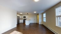 Located at 3 Chelsea in Cumberland, Maine, this single-floor residence offers a clean, modern design with an open-concept living and kitchen area that maximizes the sense of space. The kitchen is well-appointed with stainless steel appliances — including a refrigerator, range, and over-the-range microwave — white cabinetry, and a peninsula that provides additional counter space and doubles as a casual dining or workspace area. The flooring throughout is a rich, dark hardwood that adds warmth and contrast to the bright, neutral-toned walls and white trim. Natural light enters through a window in the living area, complemented by recessed and flush-mount ceiling lighting that keeps the space well-lit at any time of day. A baseboard heating unit is positioned along the exterior wall, providing efficient heating during Maine's cooler months. Interior doors lead to additional areas of the unit, and the overall layout flows openly and efficiently. The combination of contemporary finishes, functional design, and quality materials makes this a practical and attractive living space.