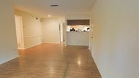 This single-floor residence offers a straightforward and functional layout designed to maximize usable living space. The main living area is expansive and open, featuring warm hardwood-style flooring that extends throughout, lending a cohesive and polished look to the interior. Neutral white walls and recessed lighting contribute to a bright, welcoming ambiance throughout the space.

A defining architectural feature is the pass-through kitchen, which opens directly into the living area via a raised countertop bar. The kitchen is equipped with light wood cabinetry and is accented by a dark window shade above, creating a subtle contrast against the otherwise light interior. This open-concept connection between the kitchen and living space supports ease of movement and interaction between the two areas.

The living area itself is generously proportioned, offering ample room for a variety of furniture arrangements. Visible HVAC vents are integrated into both the ceiling and lower wall areas, ensuring efficient climate control throughout. A white paneled door near the kitchen provides access to an adjacent area or entry point, adding to the overall connectivity of the floor plan. The space presents a clean, move-in ready canvas suited for a range of interior styles.