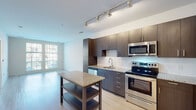 This single-floor residence showcases a thoughtfully designed open-concept layout that seamlessly connects the kitchen and living areas. The kitchen is a standout feature, outfitted with rich dark-toned cabinetry, granite countertops, a subway tile backsplash, and a full suite of stainless steel appliances including a refrigerator, electric range, built-in microwave, and dishwasher. A freestanding kitchen island with a matching granite top provides additional prep space and storage. Light hardwood flooring runs throughout the unit, lending warmth and continuity to each room. The open living area is bright and airy, benefiting from large windows that invite generous natural light and offer views of the surrounding neighborhood. The bedroom continues the light-filled aesthetic, featuring tall windows fitted with blinds and ample wall space. Storage is well-addressed with a walk-in closet equipped with wire shelving and hanging rods. The bathroom is clean and contemporary, featuring a dark wood vanity with a white countertop, a large mirror, tile flooring, and a tub-shower combination with tiled surround. Track lighting in the kitchen and clean white walls throughout contribute to a crisp, modern ambiance across the entire space.