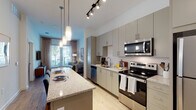 This single-floor residence is thoughtfully designed with a contemporary aesthetic that balances functionality and style throughout. The open-concept kitchen serves as a central feature, outfitted with sleek grey cabinetry extending floor to ceiling, stainless steel appliances including a range, microwave, dishwasher, and refrigerator, and a hexagonal tile backsplash that adds visual interest. A granite-topped island with pendant lighting overhead provides additional prep space and casual seating for multiple guests, making it ideal for both cooking and entertaining.

Light wood-tone flooring flows seamlessly from the kitchen into the living area, which is bright and welcoming thanks to large floor-to-ceiling windows and glass doors that open to a balcony, allowing generous natural light and outdoor views. The bedroom continues the airy theme with wide windows dressed in sheer curtains, creating a calm and restful environment.

The bathroom is well-appointed with a long granite vanity, grey cabinetry, large-format tile flooring, and direct access to a walk-in closet, adding a practical and organized element to the private quarters. Track lighting and recessed fixtures illuminate the space throughout, contributing to a clean, modern ambiance overall.