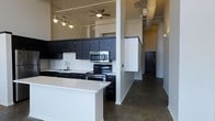 This single-floor space showcases a bold, industrial-modern aesthetic that blends contemporary finishes with raw architectural character. The open-concept kitchen serves as the centerpiece, featuring dark cabinetry that contrasts sharply with crisp white quartz countertops and a classic white subway tile backsplash. Stainless steel appliances, including a range, microwave, and dishwasher, add a sleek, functional edge to the cooking space. A kitchen island with an extended countertop provides additional workspace and casual dining potential. Overhead, a cluster of modern pendant lights illuminates the kitchen, while a ceiling fan with integrated lighting serves the adjacent living area. The soaring ceilings lend an expansive, loft-like atmosphere to the space, enhancing the sense of openness and volume. Polished concrete floors run throughout, reinforcing the industrial character and offering a durable, low-maintenance surface. Structural columns are integrated naturally into the layout, adding to the architectural interest. A wide, open corridor leads toward additional areas of the unit, with a dark-toned entry door providing a grounding contrast to the lighter walls. Overall, the space delivers a refined urban living experience with a cohesive, contemporary design language.