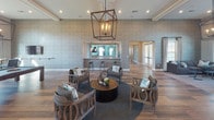 This expansive single-story space is designed with an open-concept layout that seamlessly connects multiple functional areas under one roof. Wide-plank hardwood-style flooring flows continuously throughout, unifying the various zones while contributing to a warm, cohesive aesthetic.

The kitchen serves as a central hub, featuring dark cabinetry, a generous white quartz island with an undermount sink, stainless steel appliances, and a teal glass subway tile backsplash. Pendant lighting — including elegant lantern-style fixtures — illuminates the space while adding decorative character.

A large lounge area anchors one end of the layout, highlighted by a textured accent wall, a striking geometric chandelier, and a seating arrangement centered around a round coffee table. Adjacent to this is a comfortable media lounge with a stone-clad column housing a wall-mounted television, flanked by large arched windows that invite abundant natural light and outdoor views.

The game and activity area accommodates a billiards table and multiple high-top seating clusters, enhanced by tall windows overlooking the surrounding landscape. A quieter sitting nook provides an intimate retreat with plush seating and ample natural light. Additional utility areas include a dedicated laundry station with teal tile accents and a floating countertop.