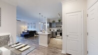 This well-appointed single-floor residence showcases a thoughtfully designed open-concept layout that seamlessly connects the kitchen, dining, and living areas. The kitchen is a standout feature, outfitted with gray shaker-style cabinetry, a white subway tile backsplash, stainless steel appliances — including a refrigerator, range, and built-in microwave — and a generous white quartz peninsula with bar seating, illuminated by modern cage-style pendant lights. The living area offers wood-look plank flooring, crown molding, a ceiling fan, and access to an outdoor area through a glass door that fills the space with natural light. A built-in shelving unit adds functional storage and visual interest near the entryway to the bedroom. The bedroom is a comfortable retreat with plush carpeting, large windows dressed in sheer curtains and plantation shutters, and ample natural light. The bathroom features gray cabinetry, a white quartz countertop with an undermount sink, and a large framed mirror, with direct access to a spacious walk-in closet equipped with multi-tier shelving and hanging storage. Throughout the residence, a cohesive neutral palette and consistent finishes create a clean, modern ambiance.