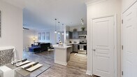 This well-appointed single-floor residence showcases a thoughtfully designed open-concept layout that seamlessly connects the kitchen, dining, and living areas. The kitchen is a standout feature, outfitted with gray shaker-style cabinetry, a white subway tile backsplash, stainless steel appliances — including a refrigerator, range, and built-in microwave — and a generous white quartz peninsula with bar seating, illuminated by modern cage-style pendant lights. The living area offers wood-look plank flooring, crown molding, a ceiling fan, and access to an outdoor area through a glass door that fills the space with natural light. A built-in shelving unit adds functional storage and visual interest near the entryway to the bedroom. The bedroom is a comfortable retreat with plush carpeting, large windows dressed in sheer curtains and plantation shutters, and ample natural light. The bathroom features gray cabinetry, a white quartz countertop with an undermount sink, and a large framed mirror, with direct access to a spacious walk-in closet equipped with multi-tier shelving and hanging storage. Throughout the residence, a cohesive neutral palette and consistent finishes create a clean, modern ambiance.