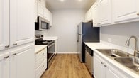 This single-story unit located in Santa Ana, California offers a well-organized layout distributed across several distinct living areas. The galley-style kitchen is equipped with stainless steel appliances, including a refrigerator, electric range, microwave, and dishwasher, complemented by white cabinetry, light stone countertops, and wood-look flooring that adds warmth to the functional space.

The living area features a combination of wood-look and carpeted flooring, neutral-toned walls, and a textured ceiling. A large window fitted with vertical blinds allows natural light to fill the room while offering views of the surrounding greenery. Adjacent rooms share a similarly neutral palette, with carpeted floors and windows that bring in outdoor views of the landscaped grounds.

The unit includes two bathrooms: one featuring a soaking tub and shower combination with a curved curtain rod, and another with a sliding glass-door shower stall. Both bathrooms are finished with white vanities, stone-look countertops, and wood-look flooring. A spacious walk-in closet with dual hanging rods and shelving provides ample storage. Wall-mounted HVAC units are present throughout, ensuring year-round climate control.