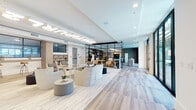 Located at 1018 N Caldwell St in Charlotte's NoDa neighborhood, this single-floor amenity space is a thoughtfully designed communal hub that blends modern aesthetics with inviting functionality. Light wood-tone flooring flows throughout, anchoring a cohesive palette of whites, warm neutrals, and charcoal accents across each distinct zone.

The main lounge area features an eye-catching illuminated ceiling installation spelling out the building's branding, serving as a bold focal point above clusters of sculptural lounge chairs and soft ottomans arranged for casual conversation. A wall-mounted fireplace and large-screen TV anchor a secondary seating area, creating a relaxed, residential atmosphere.

A glass-and-black-steel partition wall elegantly separates the game room, which houses classic arcade cabinets alongside a bistro table and seating — an entertainment alcove visible from the broader lounge. Adjacent, a generously sized communal dining area centers on a substantial reclaimed wood farm table beneath pendant lighting, accompanied by a fully equipped kitchen featuring dark cabinetry, exposed brick walls, a draft beer tap system with a stainless steel kegerator, and open shelving. Built-in banquette seating and nook areas offer quieter retreats. Floor-to-ceiling windows flood the space with natural light and provide views to the outdoor courtyard.