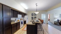 This single-floor residential unit offers a thoughtfully designed layout with a seamless flow between its main living spaces. The kitchen is a standout feature, showcasing rich dark cabinetry, granite countertops, and light hardwood flooring. A large central island with bar seating and a pendant light above serves as both a functional workspace and a casual dining area, making it ideal for shared living. White appliances and a skylight-style ceiling fixture keep the space bright and welcoming.

Adjacent to the kitchen, the living area provides generous room for relaxing and entertaining, with soft carpet underfoot and light-toned walls that create a calm, open atmosphere. Hardwood flooring in the common areas transitions cleanly into carpeted spaces throughout.

The unit includes multiple bedrooms, each featuring plush carpet, natural light from windows dressed with bold curtains, ceiling fans, and ample room for a bed and desk setup. Each room offers a comfortable, private retreat.

Two full bathrooms mirror each other in design, featuring slate-style tile flooring, dark vanity cabinetry with granite countertops, mirrored medicine cabinets, and shower-tub combinations. The consistent finishes throughout the unit give it a cohesive and well-maintained appearance, making it a practical and comfortable living environment.