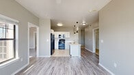This single-floor residence offers a thoughtfully designed layout encompassing two bedrooms, two bathrooms, a kitchen, and a dedicated closet. The open-concept kitchen serves as a central hub, featuring white cabinetry, marble-look countertops with a double stainless steel sink, black appliances including a refrigerator, range, and microwave, and light tile flooring. A peninsula with pendant lighting connects the kitchen to the living area, which is finished with light wood-look flooring throughout. The living and dining area is bright and airy, highlighted by a bank of large windows that frame outdoor greenery views and allow generous natural light to fill the space. A ceiling fan adds both function and comfort. The primary bedroom is a well-proportioned retreat with dual windows, a ceiling fan, and the same wood-look flooring. It is complemented by a dedicated walk-in closet equipped with wire shelving and hanging rods. The two bathrooms feature light tile flooring, vanities with mirrors, and clean, functional layouts. One bathroom includes a warm wood-toned vanity, while the second offers a bold, framed mirror as a design accent. Neutral gray tones throughout the unit create a cohesive, modern aesthetic.