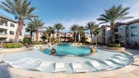 Located at 12325 Shadow Creek Pkwy in Pearland, TX, this single-floor clubhouse amenity space offers a well-appointed community hub designed for both relaxation and recreation. The centerpiece of the outdoor area is a stunning resort-style pool surrounded by tall palm trees, decorative sphere water features, in-pool loungers, and a spacious sun deck — all set beneath the wide Texas sky. Inside, the clubhouse is thoughtfully designed with warm, contemporary finishes throughout. The lounge area features rich hardwood-style flooring, patterned chevron upholstered seating, and an eclectic cluster of Edison bulb pendant lights that lend a modern-industrial ambiance. Large arched windows and glass doors frame views of a landscaped courtyard with outdoor seating, seamlessly connecting the interior to the exterior. The game room is a standout feature, showcasing a full-size billiards table, a shuffleboard table, and a wall-mounted flat-screen TV set against a dark wood accent wall. A tray ceiling with warm accent lighting adds architectural depth to the space. An adjacent dining and bar area provides additional seating and workspace. Together, these spaces create an engaging and social environment tailored for community living.