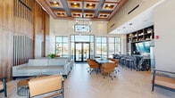 This thoughtfully designed amenity space spans a generous single floor, offering a diverse range of functional areas finished to a high standard. The centerpiece is a dramatic living room with soaring double-height ceilings adorned with an elegant coffered wood ceiling detail and a statement mid-century modern chandelier. Floor-to-ceiling black-framed windows and glass doors flood the space with natural light while providing views of an outdoor pool area. Herringbone-patterned tile flooring, a textured wood slat accent wall, and a built-in bar with dark cabinetry and display shelving contribute to the room's sophisticated, contemporary ambiance.

Adjacent to the living room is a dedicated exercise room equipped with treadmills, ellipticals, weight machines, and a dumbbell rack. Large black-framed windows line the gym walls, offering natural light and exterior views, while full-length mirrors and track lighting complete the functional setup.

The bedroom is a bright, simply appointed room featuring herringbone tile flooring and a large window overlooking an outdoor courtyard with palm trees. A glass-enclosed private office pod with black steel framing sits nearby in the hallway area, offering a focused workspace within the open layout.

The bathroom is finished with full marble-look tile on both the floors and walls, complemented by a dark granite vanity countertop with wood cabinetry and accessible grab bars.