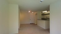 This single-floor space offers a straightforward, functional layout designed for comfortable everyday living. The main living area is generously sized, finished with wall-to-wall plush carpeting in a warm neutral tone that extends throughout the open floor plan. Smooth, cream-colored walls and standard-height ceilings create a clean, understated backdrop, while overhead lighting provides consistent illumination throughout the space.

Positioned to one side of the living area, the kitchen is partially open to the main room and separated by a raised breakfast bar with a dark countertop, offering a casual dining or workspace option. The kitchen features light-toned cabinetry, built-in appliances including a microwave, and under-cabinet fluorescent lighting that brightens the work surfaces. The transition between the carpeted living area and the kitchen flooring is clearly defined, adding a practical distinction between zones.

A white paneled door is visible at the far end of the living space, indicating access to an additional room or entry point. The overall aesthetic is neutral and move-in ready, with a simple, cohesive palette suited to a variety of furnishing styles.