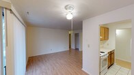 This single-floor space offers a straightforward, functional layout centered around an open living area. The room features light-toned hardwood-style flooring that runs throughout, contributing to a bright and clean aesthetic. A ceiling fan with an integrated light fixture is centrally mounted, providing both ventilation and ambient lighting. The walls are finished in a neutral white, complemented by a textured popcorn ceiling. A doorway at the far end of the living area leads to what appears to be a hallway with access to additional rooms, including a space that resembles a closet or storage area. To the right, the kitchen is partially visible, showcasing light wood cabinetry and a white range, suggesting a compact but practical cooking space. The open sightlines between the living area and kitchen create a connected feel. Overall, the space presents a modest, no-frills interior with a neutral palette and practical finishes suited for everyday living.