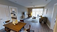 This single-floor residence features an open-concept layout that seamlessly connects the dining, living, and sleeping areas. The space is finished with wide-plank hardwood-style flooring throughout, providing a warm and cohesive aesthetic. A ceiling fan overhead adds both functionality and a contemporary touch to the main living area.

The dining area is anchored by a solid wood table with mixed seating, complemented by decorative accents including ceramic vessels in earthy tones. The living room is generously proportioned, centered around a neutral sofa accented with mustard-yellow throw pillows and a wood-framed coffee table. A cozy accent chair in warm brown tones adds character alongside a mid-century-inspired media console and wall-mounted television.

Large floor-to-ceiling sliding glass doors dominate one wall, allowing abundant natural light to pour in and offering access to an outdoor balcony area. A piece of abstract wall art introduces a pop of color to the otherwise neutral palette. The bedroom, visible through an open doorway, continues the modern aesthetic with layered textiles and consistent flooring, contributing to the space's overall sense of flow and cohesion.