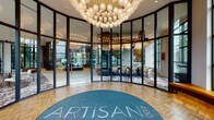 Artisan 400's clubhouse is a single-level amenity space designed with a sophisticated, contemporary aesthetic. The entrance rotunda immediately establishes the tone, featuring a curved glass-and-steel curtain wall and an oversized sculptural chandelier composed of clustered globe pendants, with herringbone-patterned wood flooring and a branded welcome mat beneath.

Beyond the entry, the main lounge unfolds as a generously scaled gathering space anchored by a dramatic floor-to-ceiling dark stone fireplace. Seating arrangements of curved lounge chairs and modular sofas surround the hearth, all positioned atop a bold, richly patterned area rug with swirling earth tones. Coffered ceilings with warm integrated lighting add architectural depth overhead, while geometric gold-accented room dividers define zones without fully enclosing them.

A conference and dining area features a large wood communal table, sculptural sputnik-style chandelier, and full-height steel-framed windows offering outdoor courtyard views. A kitchenette equipped with stainless steel appliances, including a refrigerator and dishwasher, supports entertaining. A game area with a billiards table, a dedicated package room, and additional lounge seating round out the well-appointed community space.