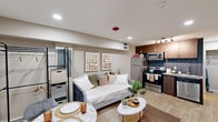 Located at 9220 N IH-35 Service Rd in Austin, TX, this thoughtfully designed unit maximizes every square foot of its compact footprint across a single floor. The open-plan layout seamlessly integrates the living, dining, and kitchen areas into one cohesive space, creating a functional and inviting environment.

The living area features light wood-look flooring that runs throughout, recessed ceiling lighting, and neutral-toned walls that contribute to a bright, airy feel. A cozy seating arrangement pairs well with a round coffee table, while a compact dining set near the entryway offers a dedicated space for meals.

The kitchen is a standout feature, equipped with a full suite of stainless steel appliances — including a refrigerator, electric range, microwave, and dishwasher. Rich walnut-toned cabinetry with gold hardware is complemented by a striking navy blue subway tile backsplash, adding a modern, stylish contrast. Track lighting above the kitchen provides focused illumination for the workspace.

The bathroom continues the cohesive design aesthetic, featuring a vessel sink atop a walnut-toned vanity with a stone countertop, wood-look flooring, and a shower-tub combination. Clean lines and warm tones give the bathroom a polished, contemporary feel. Overall, the unit delivers a well-appointed, modern living experience in a compact and efficient layout.