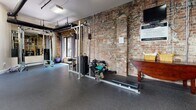 This fitness facility seamlessly blends historic architectural character with modern gym functionality. The space is defined by striking exposed brick walls that lend a raw, industrial warmth, complemented by original heavy timber support columns that speak to the building's heritage. Durable rubber flooring — in both dark speckled and blue speckled varieties across different zones — provides a safe, practical surface throughout the workout areas.

The gym is well-appointed with a comprehensive selection of equipment to support a full-body fitness routine. Cardio options include treadmills and elliptical cross-trainers, while the strength training area features a cable/weight machine, a leg press machine, a adjustable weight bench, and a selection of dumbbells. A stability ball and additional free weights round out the offerings for functional and flexibility training.

Large wall-mounted mirrors amplify the sense of space and allow for proper form checks during workouts. Overhead fluorescent lighting ensures the facility is well-lit throughout. Exposed pipes and ductwork along the ceiling contribute to the space's industrial aesthetic. The facility is accessed through a sleek black-framed glass door, and a clearly marked exit sign ensures safety compliance. Overall, the space offers a functional and characterful environment well-suited for a comprehensive fitness regimen.