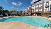 Located at 9240 Brukhaus in Raleigh, NC, The Exchange is a well-appointed residential community featuring a thoughtfully designed single-level amenity area that serves as its social hub. The covered breezeway entrance is framed by arched ceilings, shiplap-accented walls, and a branded runner mat bearing "The Exchange," creating a polished first impression. Built-in water fountains and utility access points, including a dedicated fire riser room, reflect the space's practical infrastructure.

At the heart of the outdoor area is a generously sized, freeform resort-style swimming pool with a glistening turquoise surface, surrounded by a broad concrete pool deck ideal for lounging. Ample seating with colorful umbrellas lines the perimeter, providing shade and comfort. A wood pergola structure anchors one side of the pool area, offering a shaded transitional space between the building's entrance and the pool deck.

Adjacent to the pool, a landscaped outdoor lounge area features wicker furniture with vibrant green cushions, mature trees, and a wall-mounted flat-screen television set within a brick surround — perfect for outdoor entertaining. Multi-story residential buildings with large windows and private balconies frame the complex, creating a cohesive and attractive architectural backdrop throughout.
