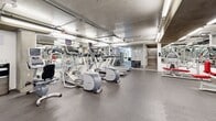 Located at 801 N Fairfax in Los Angeles, this well-equipped fitness center offers a comprehensive exercise environment across a single open floor. The space is designed for functionality, featuring durable rubber tile flooring that covers the entire workout area and provides both comfort and safety during exercise. Overhead fluorescent strip lighting illuminates the room evenly, ensuring excellent visibility throughout.

The main exercise floor is lined with floor-to-ceiling mirrored walls that visually expand the space and allow users to monitor their form. A robust selection of cardio equipment is available, including treadmills, elliptical trainers, and a stair climber, all positioned along the mirrored walls and accompanied by mounted flat-screen televisions. The strength training section features a variety of selectorized weight machines with red upholstered pads, a dumbbell rack with a full range of free weights, and kettlebells for functional training.

Exposed concrete columns and visible overhead infrastructure give the space an industrial aesthetic, complemented by crisp white walls. A glass-enclosed lounge or reception area is accessible from the main floor, featuring decorative patterned wall accents, seating, and a welcoming atmosphere. The combination of diverse equipment, thoughtful layout, and modern design elements makes this fitness center a comprehensive facility for a full-body workout.