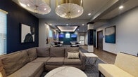 Located at 1998 Persimmon Way in Tipp City, Ohio, this single-floor community clubhouse offers a thoughtfully designed gathering space with a sophisticated, modern aesthetic. The grand entrance lobby commands attention with soaring double-height ceilings, large decorative lattice shelving units flanking the entry doors, and bold pops of yellow in the furnishings that contrast beautifully against the neutral tile flooring. A striking floor-to-ceiling stacked stone fireplace anchors the main lounge area, complemented by a wall-mounted television and warm tile flooring that flows seamlessly throughout. The lounge features plush sectional seating, contemporary accent chairs, geometric glass coffee tables, and curated wall art, creating an inviting atmosphere for relaxation and socializing. Overhead, statement pendant light fixtures and coffered ceiling details add a refined layer of elegance. The kitchen and dining area showcases navy cabinetry, a large quartz-topped island with bar seating, stainless steel appliances, pendant lighting, and a built-in banquette with golden-yellow upholstery set against deep navy walls. Additional entertainment spaces, visible in the layout, include a billiards table and multiple television viewing areas with casual seating. The space balances upscale design with functional communal living throughout.