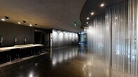 This sophisticated lobby space presents a bold, contemporary architectural design that immediately commands attention. The single-floor layout offers a spacious, open-plan environment defined by a refined dark palette and high-end material choices throughout.

The polished large-format dark tile flooring reflects the carefully positioned track and recessed lighting overhead, creating a dramatic interplay of light and shadow across the space. A striking curved ceiling element adds sculptural dimension, while the exposed concrete-finish ceiling in key zones reinforces the industrial-luxe aesthetic.

A dominant feature is the expansive backlit metallic wall panel, its burnished steel surface illuminated by a row of directional spotlights that accentuate its texture and depth. Complementing this is a series of floor-to-ceiling sheer glass or translucent chain curtain partitions, which serve as elegant spatial dividers while adding a sense of layered transparency.

Near the entrance, tall narrow windows allow natural light to filter in, framing views of the exterior greenery. A built-in reception counter or display table with bar-height seating anchors one end of the space, while a bank of mailboxes and a seating area are neatly integrated along one wall. The overall ambiance is sleek, refined, and architecturally distinctive.