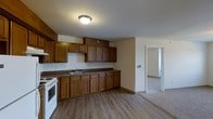 This single-floor residence offers a practical and straightforward layout designed for comfortable everyday living. The kitchen serves as a central feature of the home, showcasing warm, dark-toned wood cabinetry both above and below the countertops, providing generous storage throughout. A double-basin sink is positioned along the main counter run, accompanied by a white range, making the kitchen both functional and classic in style. The dark laminate countertops complement the rich wood tones of the cabinetry. Light wood-look vinyl plank flooring runs through the kitchen area, offering durability and a clean, neutral aesthetic. The open layout allows for a smooth transition from the kitchen into the adjacent living area, which features carpeted flooring. A doorway visible from the kitchen leads further into the home, connecting to additional rooms and suggesting a well-organized, flowing floor plan. Flush-mount ceiling lighting provides consistent illumination throughout. The overall ambiance is clean, neutral, and move-in ready, with a traditional design sensibility that prioritizes functionality and practicality.