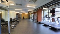 This fitness facility offers a generously sized, open workout floor designed to accommodate a wide range of exercise routines. The space is finished with durable rubber flooring in a dark speckled tone, providing both safety and comfort underfoot. Overhead, a drop-panel ceiling houses evenly distributed fluorescent lighting, ensuring the entire area is well-lit throughout. Exposed industrial ductwork runs along one side of the ceiling, adding a utilitarian aesthetic to the space.

The strength training section lines one wall and features a variety of cable machines, weight racks, and adjustable benches, offering ample options for resistance-based workouts. Full-length mirrors are positioned along one side, supporting proper form during exercise. On the opposite side of the floor, cardio equipment including treadmills and rowing machines is arranged near large windows that allow natural light to filter in and provide views to the exterior. Accent walls in warm terracotta and golden yellow tones add visual warmth to the otherwise neutral environment. The open layout ensures comfortable circulation throughout, making the space functional for both individual training and busier periods of use.