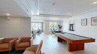 Located at 700 City View Loop in Midlothian, VA, this single-floor community clubhouse offers a well-appointed and versatile gathering space designed with a modern aesthetic. Light hardwood flooring runs throughout, complemented by crisp white walls, crown molding, and coffered ceiling details that lend an air of refined elegance.

The main lounge area centers around a wall-mounted flat-screen television framed by classic wainscoting, with plush sectional seating arranged over a deep teal area rug. Large windows and a set of double glass doors flood the room with natural light and provide direct views of the outdoor pool area. An adjacent seating nook features wicker chairs gathered around a petite bistro table beneath a softly lit tray ceiling.

A full-size billiards table anchors the game room, accompanied by comfortable lounge chairs and additional seating for spectators. The kitchen is a standout feature, showcasing floor-to-ceiling powder-blue subway tile, white shaker cabinetry, a stainless steel refrigerator, and a spacious white island with bar seating beneath sculptural pendant lights. A separate dining alcove provides banquette-style seating with warm amber upholstery and round white tables, ideal for casual gatherings. Together, these spaces create a functional, inviting environment suited for socializing and community events.