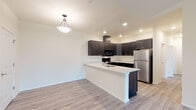 This single-floor residence offers a thoughtfully designed open-concept layout that seamlessly connects the kitchen and living areas. The kitchen is a standout feature, equipped with dark espresso-toned shaker-style cabinetry, crisp white countertops, stainless steel appliances including a full-size refrigerator, a black range with overhead microwave, and a built-in dishwasher. A peninsula with an integrated sink and pendant lighting above serves as a natural divider between the kitchen and the adjoining living space. Light wood-style flooring runs through the kitchen and transitional areas, while plush carpet covers the living room and bedrooms, adding warmth underfoot. The living area opens directly to a private balcony through sliding glass doors, offering views of surrounding greenery. A dedicated walk-in pantry or storage closet provides ample organizational space near the kitchen. The primary bathroom features a long dual-sink vanity with dark cabinetry and a wide mirror, while a secondary bathroom includes a full tub-shower combination. Bedrooms are carpeted and receive natural light through large windows with wooded outdoor views. Recessed lighting and neutral wall tones throughout the space create a clean, modern, and welcoming ambiance.