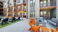 Located at 1601 Overbrook Rd in Richmond, Virginia, this outdoor amenity space serves as a central courtyard for a contemporary multi-story residential community. The building exterior showcases a modern mixed-material design, combining classic red brick with board-and-batten siding in neutral gray and white tones, complemented by warm wood-accented balconies and metal railings throughout.

The courtyard is designed for both social gatherings and relaxed outdoor living. A dedicated grilling station features multiple stainless steel gas grills built into a sleek counter, accompanied by a high-top communal table with bold red bar seating and a yellow market umbrella, making it an ideal space for outdoor dining and entertaining.

Adjacent to the grill area, a comfortable lounge zone is anchored by a built-in brick fire pit with a stone surround, encircled by vibrant orange Adirondack chairs and modern upholstered outdoor sofas. A turf lounging area with chaise seating adds a casual, recreational element to the space.

Private ground-level entries with individual metal railings and fenced patios are integrated into the courtyard design, offering direct outdoor access. The entire space is finished with large-format light gray pavers, providing a clean and cohesive aesthetic throughout. The open-air layout allows for ample natural light and views of the surrounding building architecture.