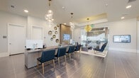 This single-story space functions as a sophisticated leasing or welcome center, thoughtfully designed to make a strong first impression. The layout is open and flowing, anchored by a long reception desk finished in a warm wood-tone veneer, accompanied by a row of navy blue upholstered chairs with gold-toned legs — ideal for one-on-one consultations.

The main area transitions seamlessly into a welcoming lounge, where curated seating arrangements feature plush chairs in charcoal leather and velvet with gold metal frames, grouped around low coffee tables atop neutral area rugs. Large-format dark tile flooring with subtle linear detailing runs throughout, contrasting beautifully with the crisp white walls.

Decorative elements elevate the space, including cascading glass globe pendant lights, a starburst gold chandelier, and a wall of metallic disc sculptures flanking a vertical slatted wood accent panel housing a flat-screen display. A glass-walled private office with brown leather seating provides a discreet consultation area.

Floor-to-ceiling windows and glass partitions invite abundant natural light, connecting the interior to an adjoining lounge or amenity space beyond. The overall ambiance is polished, contemporary, and professionally inviting.