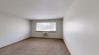 This single-floor unit features a practical and well-organized layout encompassing two bedrooms, two bathrooms, a living room, a kitchen, a closet, and two connecting hallways. The living room is a generous, open area finished with neutral-toned carpet, warm wood baseboard trim, and light gray walls, providing a clean and versatile backdrop. It includes a thermostat and multiple electrical outlets. The kitchen is equipped with rich, dark reddish-brown cabinetry, a gas range, light-toned countertops, and tile-style flooring, offering a functional workspace. The spacious primary bedroom features carpet flooring, a large picture window with an under-window air conditioning unit and baseboard heating, allowing for natural light and outdoor views. The second bedroom is similarly carpeted with a window fitted with blinds and baseboard heating. A walk-in closet with dual hanging rods and shelving provides ample storage. The two bathrooms include one with classic white mosaic tile flooring and a full tub-shower surround. Throughout the unit, warm wood doors and trim provide consistent detailing, while the neutral color palette and carpeted living areas create a cohesive and functional residential environment.
