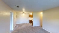 This single-floor residence offers a straightforward and functional layout designed for comfortable everyday living. The main living area is generously sized, featuring neutral-toned carpet flooring that extends throughout the space, creating a warm and cohesive feel underfoot. A ceiling fan with an integrated light fixture is centrally mounted, providing both air circulation and ambient lighting. The textured popcorn ceiling adds a classic touch to the overall aesthetic.

The living area flows seamlessly toward the kitchen, which is partially separated by a half-wall partition, offering a degree of definition between spaces while maintaining an open feel. The kitchen is equipped with warm honey-toned wood cabinetry providing ample storage, light-colored countertops, and a deep-toned base cabinet housing the sink area. A fire extinguisher is visibly mounted, reflecting attention to safety.

The walls throughout are painted in a soft, neutral tone, contributing to a bright and airy atmosphere. Visible HVAC venting in the ceiling indicates a central air system. A doorway on the far left leads to additional rooms, suggesting further functional spaces beyond what is immediately visible. The overall layout is clean, practical, and well-suited for efficient day-to-day use.