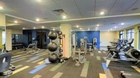 This fitness center offers a spacious, single-floor layout designed to accommodate a wide range of workout styles. The open floor plan provides ample room for movement between equipment stations, with a recessed drop ceiling fitted with evenly distributed recessed lighting that ensures the space is well-lit throughout. A bold blue accent wall serves as a defining visual element, adding energy and vibrancy to the environment.

The flooring features a multi-toned striped carpet with blue, green, and gray tones, creating clear zones within the space while adding a modern aesthetic. The strength training area is well-stocked with cable machines, weight benches, and a variety of resistance equipment positioned throughout the room. A dedicated functional training zone houses a full rack of stability balls in multiple sizes, medicine balls, resistance bands, and a BOSU balance trainer, catering to dynamic and core-focused workouts.

A structural column near the center of the space subtly divides workout zones without disrupting the overall open feel. Large glass doors or windows near one wall hint at additional adjoining areas, contributing to an expansive, connected atmosphere well-suited for a comprehensive fitness facility.