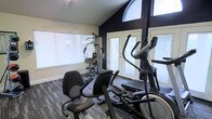 This dedicated fitness space is thoughtfully designed to support a well-rounded workout routine. The room is outfitted with a selection of exercise equipment, including a multi-function strength training station, an elliptical trainer, and a stationary recumbent bike, providing options for both cardiovascular and resistance training. 

Natural light fills the space through a combination of large windows fitted with horizontal blinds and a prominent arched window positioned near the vaulted ceiling, adding an architectural focal point while brightening the interior. A set of double glass-panel doors opens to the exterior, enhancing ventilation and further contributing to the room's airy feel.

The design aesthetic is clean and purposeful, with a bold dark accent wall contrasting against lighter surrounding walls to create a dynamic, motivating atmosphere. Durable patterned carpet flooring runs throughout, offering both comfort underfoot and a practical surface for a fitness environment. The angled ceiling adds height and an open sense of volume to the space, making it feel more expansive than its footprint might suggest. Overall, this fitness room combines functional equipment, strong natural light, and intentional design elements to create an inviting and effective workout environment.