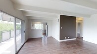This single-floor space offers a clean, modern aesthetic defined by an open-concept layout that connects the living area to the kitchen. The flooring throughout is a warm wood-look laminate that adds a sense of warmth and continuity across the space. The living area is characterized by a high textured ceiling with an exposed beam detail, enhancing the sense of volume and architectural interest. A striking dark brown accent wall provides a bold contrast against the otherwise neutral, light gray and white color palette, adding depth and character to the room. A sliding glass door on one side of the living area allows for natural light to filter in while offering convenient access to the exterior. The kitchen is visible from the main living space, separated by a pass-through window that promotes an open, connected feel. The kitchen is equipped with stainless steel appliances, including a refrigerator and range. Natural light enters through a window above the kitchen area, keeping the space bright and welcoming. White baseboards and trim throughout provide a crisp, finished look that ties the interior design together cohesively. The overall ambiance is airy, minimal, and move-in ready.