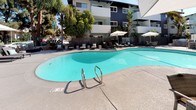 Located at 425 Merrimac Way in Costa Mesa, California, this outdoor amenity space offers a well-appointed resort-style retreat designed for relaxation and leisure. At its centerpiece is a generously sized freeform swimming pool with a striking turquoise finish, equipped with stainless steel entry rails and surrounded by a smooth, spacious concrete deck. Adjacent to the pool sits a dedicated spa, providing a quieter space for unwinding alongside the main pool area.

The deck is thoughtfully furnished with contemporary padded chaise lounges accented by light blue throw pillows, each positioned beneath large, modern white market umbrellas for ample shade. Additional outdoor dining and seating areas feature wicker chairs and glass-top tables, also shaded by umbrellas, making the space equally suitable for casual outdoor meals or socializing.

Privacy screens and wrought iron fencing define the boundaries of the area, offering seclusion while maintaining an open, airy atmosphere. Mature palms, tropical plantings, and ornamental shrubs frame the perimeter, contributing to the lush, Southern California ambiance. A covered cabana-style structure in the background adds a dedicated lounge zone, enhancing the overall functionality of this outdoor space. The sunny Costa Mesa climate makes this area an ideal year-round outdoor destination.