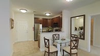 This single-floor residence offers a thoughtfully designed layout that blends functionality with classic elegance. The kitchen is a standout feature, showcasing rich dark wood cabinetry that extends to the ceiling, providing ample storage space. Stainless steel appliances, including a full-size refrigerator and built-in microwave, add a modern touch, while granite countertops lend a refined, polished aesthetic. A raised breakfast bar-style peninsula serves as a natural transition between the kitchen and the adjacent dining area, making the space ideal for both everyday living and entertaining. The dining area comfortably accommodates a glass-top table with classic cross-back wooden chairs, creating an inviting atmosphere for meals. Light-toned tile flooring runs throughout, contributing to a clean, airy feel and complementing the warm tones of the cabinetry. Flush-mount ceiling light fixtures provide consistent, well-distributed illumination across the space. A large mirror near the dining area enhances the sense of openness, reflecting light and visually expanding the room. The front entry door is conveniently situated near the kitchen, offering a practical and welcoming flow into the home. Overall, the space presents a cohesive, warm, and functional interior with timeless design details.