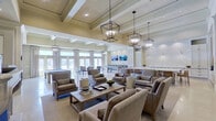 This refined single-floor amenity space is designed with a cohesive, upscale aesthetic that blends transitional architecture with comfortable, hospitality-inspired interiors. The open layout is anchored by soaring coffered ceilings adorned with striking geometric lantern-style pendant fixtures that cast warm, inviting light throughout. Polished travertine flooring runs continuously across the space, enhancing its sense of openness and grandeur.

The main lounge area features multiple groupings of tailored upholstered armchairs and sofas in neutral tones, centered around a wood coffee table, creating an ideal setting for relaxed gatherings. A classic fireplace with a refined mantel serves as a focal point in an adjacent seating area, flanked by French doors that invite abundant natural light. Framed artwork and built-in wall sconces add curated detail to the walls.

The game room accommodates a full-sized billiard table alongside additional gaming surfaces, all bathed in natural light from a wall of French doors and floor-to-ceiling drapery panels. A dedicated catering kitchen showcases floor-to-ceiling white cabinetry, marble countertops, a decorative patterned tile backsplash, a large central island with barstool seating, and a coffee station, making it highly functional for events and social gatherings.
